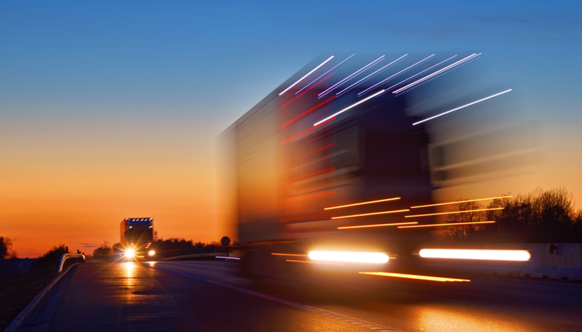 A truck moves along a highway at sunset, with the sky ablaze in shades of orange and pink, creating a picturesque scene.