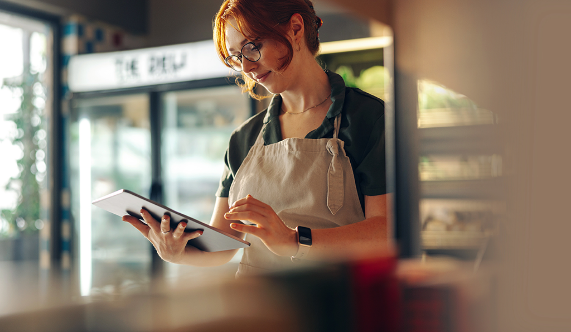 In a deli, a woman wearing an apron is engaged with a tablet, likely managing orders or tasks.